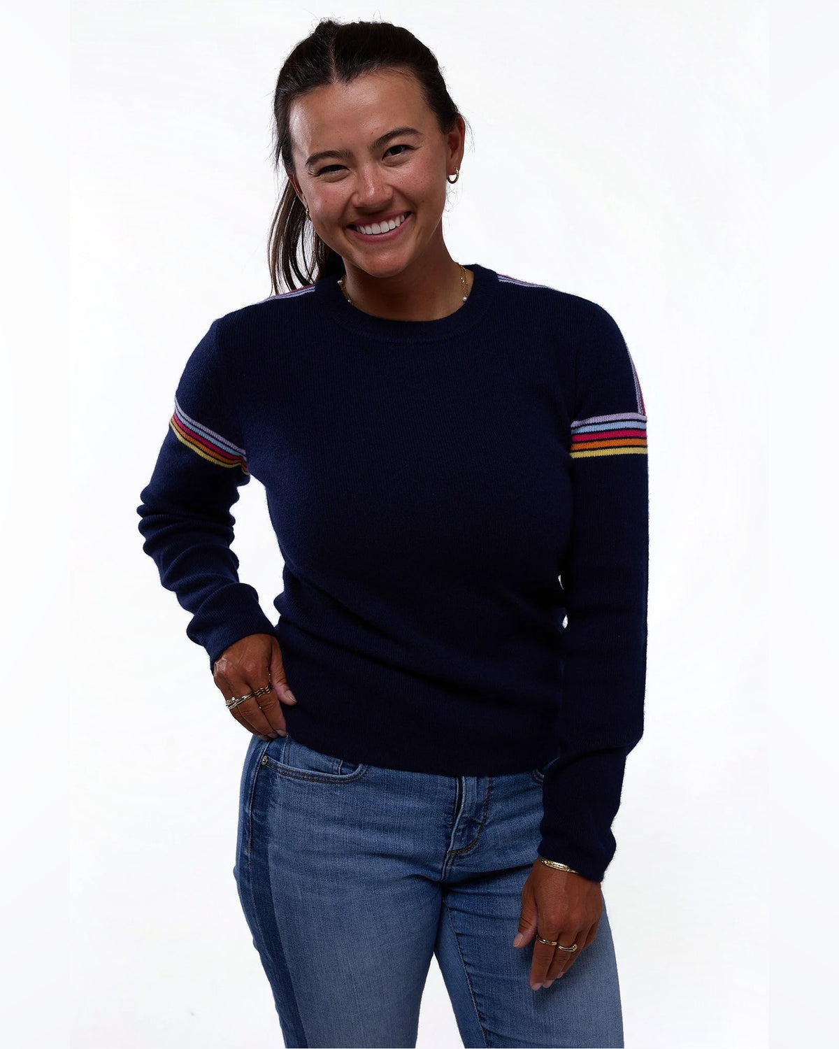 A smiling woman wearing a navy blue crewneck sweater with colorful striped details on the sleeves, paired with blue jeans, against a plain white background. - Midnight