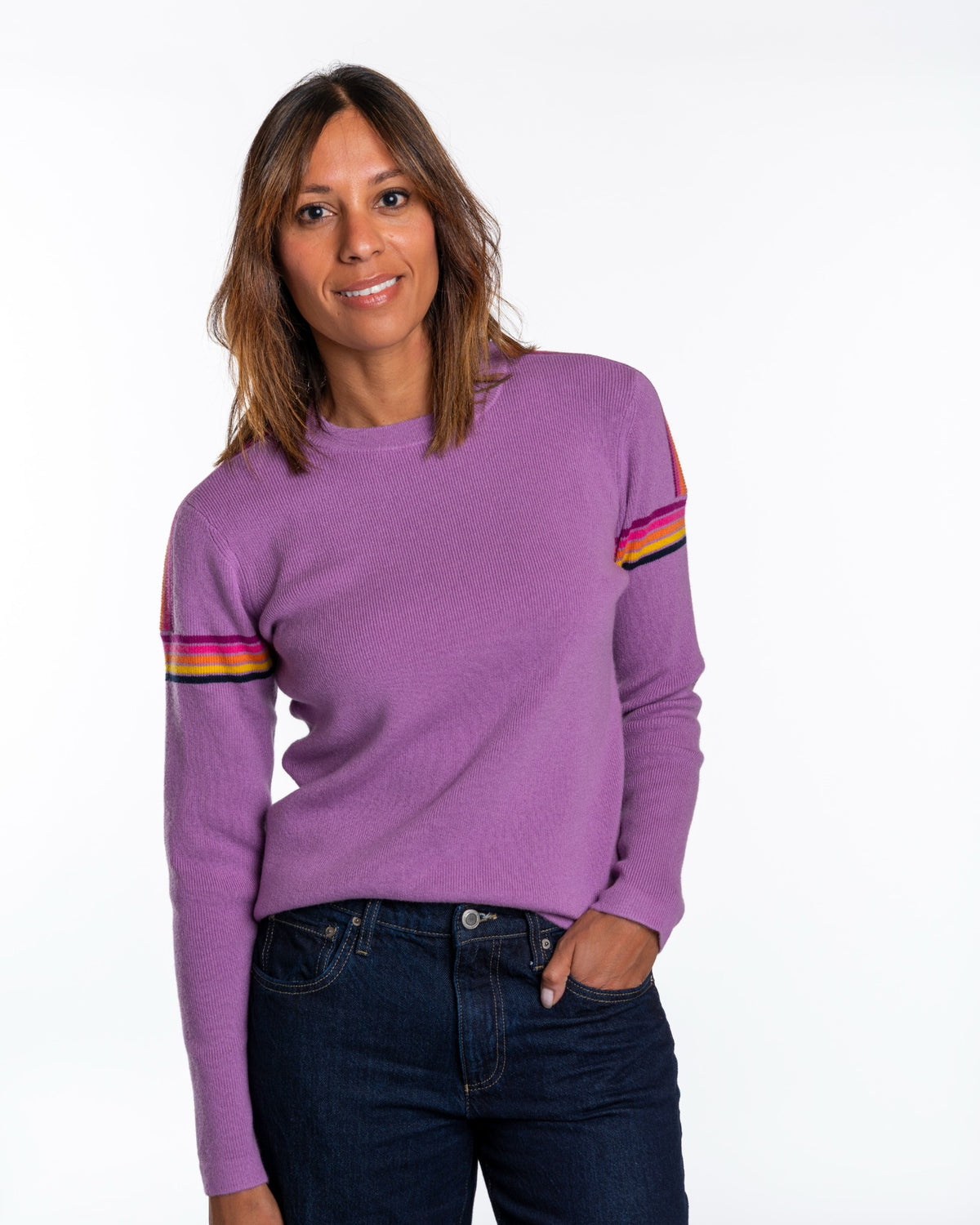 A woman wearing a purple crewneck sweater with colorful striped details on the sleeves, paired with dark blue jeans, against a plain white background. - Alpenglow