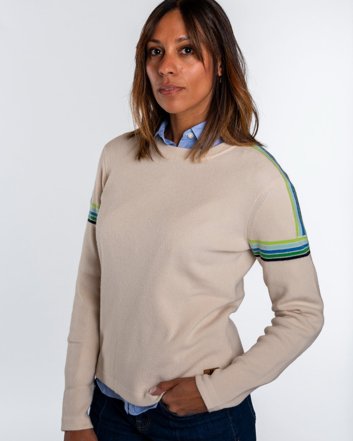 A woman wearing a beige crewneck sweater with striped accents on the sleeves, layered over a light blue collared shirt and dark blue jeans, against a plain light gray background. - Snow