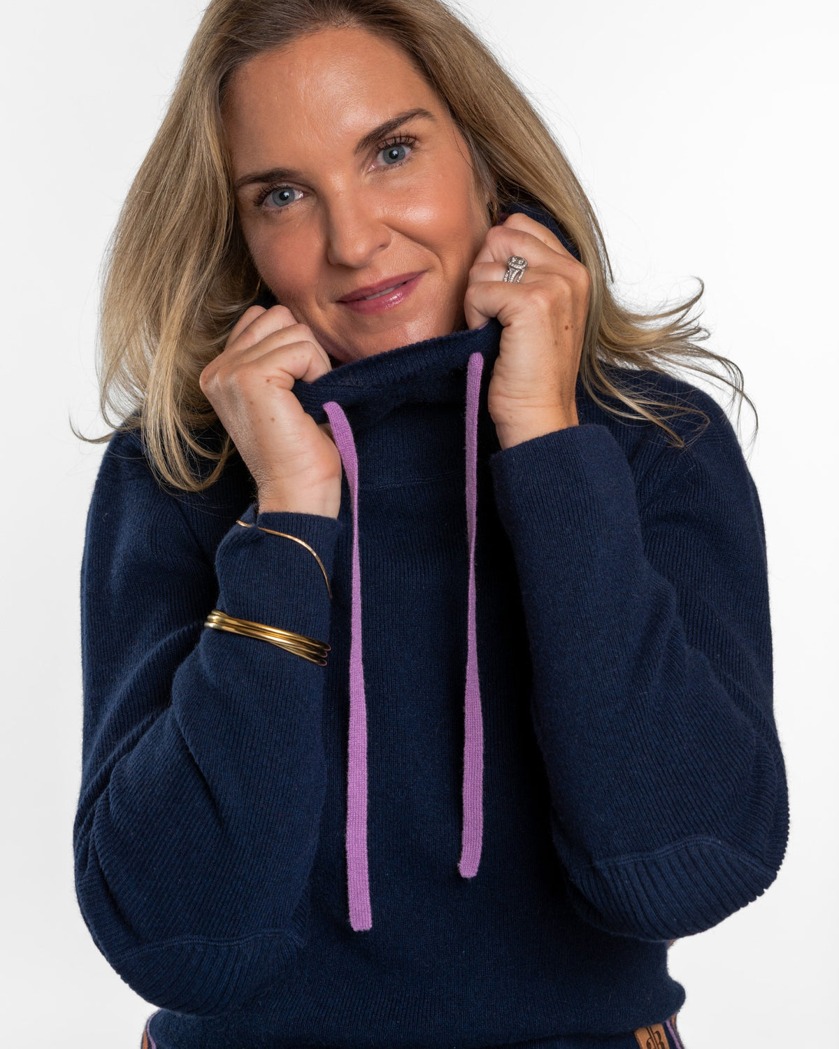 A woman wearing a navy blue knit sweater with lavender drawstrings, holding the collar open. She is wearing gold bracelets on one wrist and a ring on her finger, set against a plain white background. - Navy