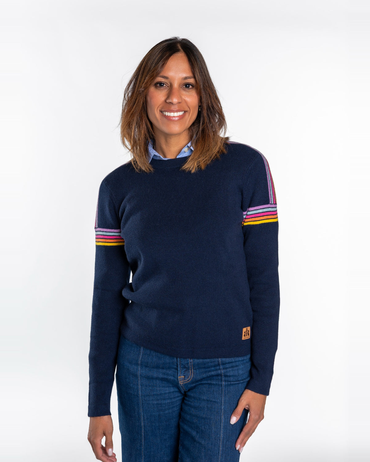 A woman wearing a navy blue crewneck sweater with colorful striped details on the sleeves, paired with blue jeans, against a plain white studio background. - Navy