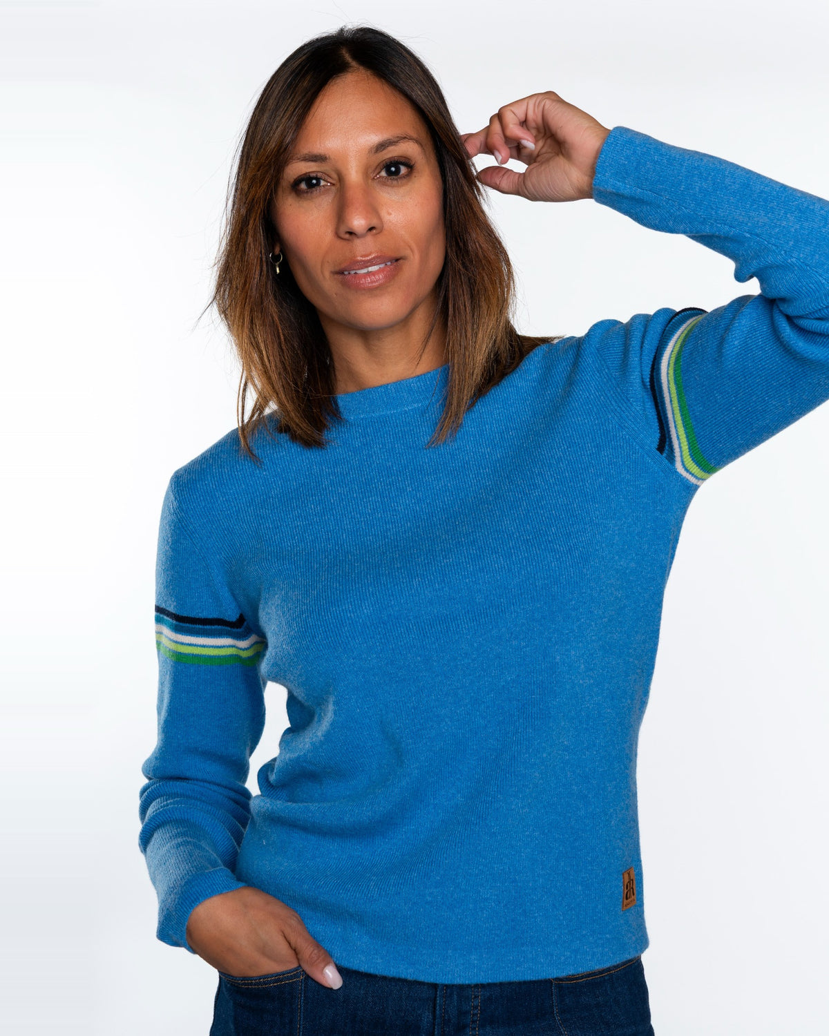 A woman wearing a bright blue crewneck sweater with striped details on the sleeves, paired with dark blue jeans, against a plain white studio background. - Sky
