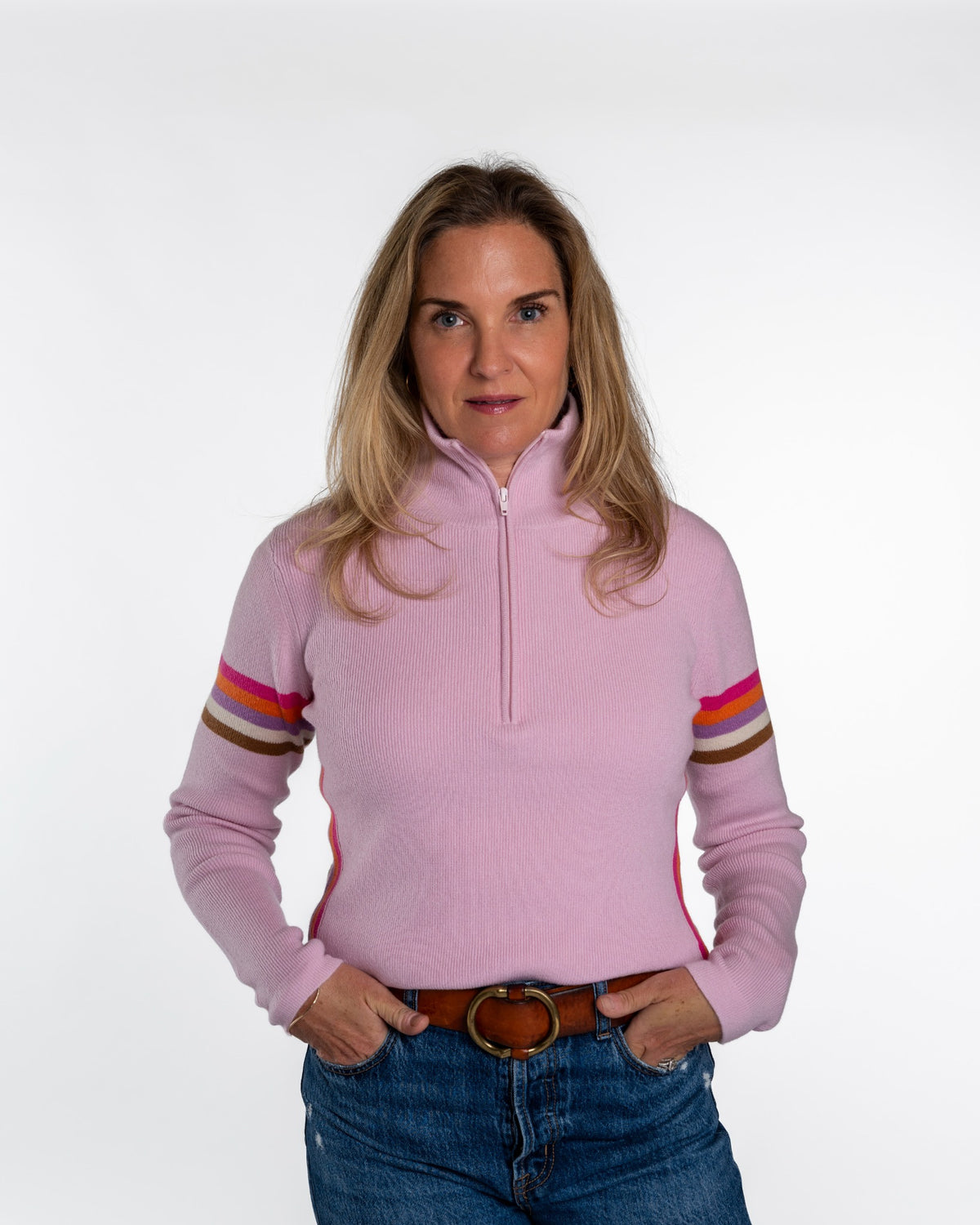 A woman modeling a light pink quarter-zip long-sleeve sweater with colorful striped details on the sleeves, paired with blue jeans and a brown belt, against a plain white studio background. - Rose