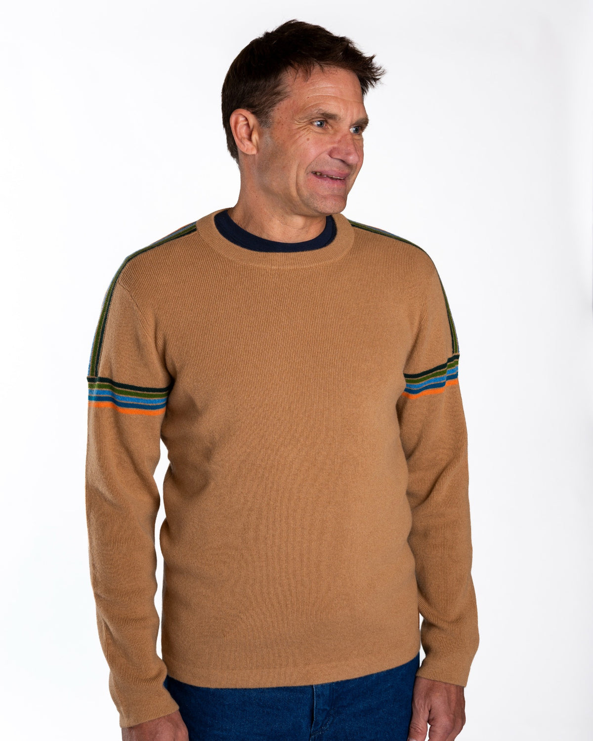 A man wearing a tan ribbed knit sweater with dark navy trim at the crew neck and sleeve cuffs, featuring thin green, blue, and orange stripes near the upper sleeves, paired with blue jeans against a plain white background.  - Sand