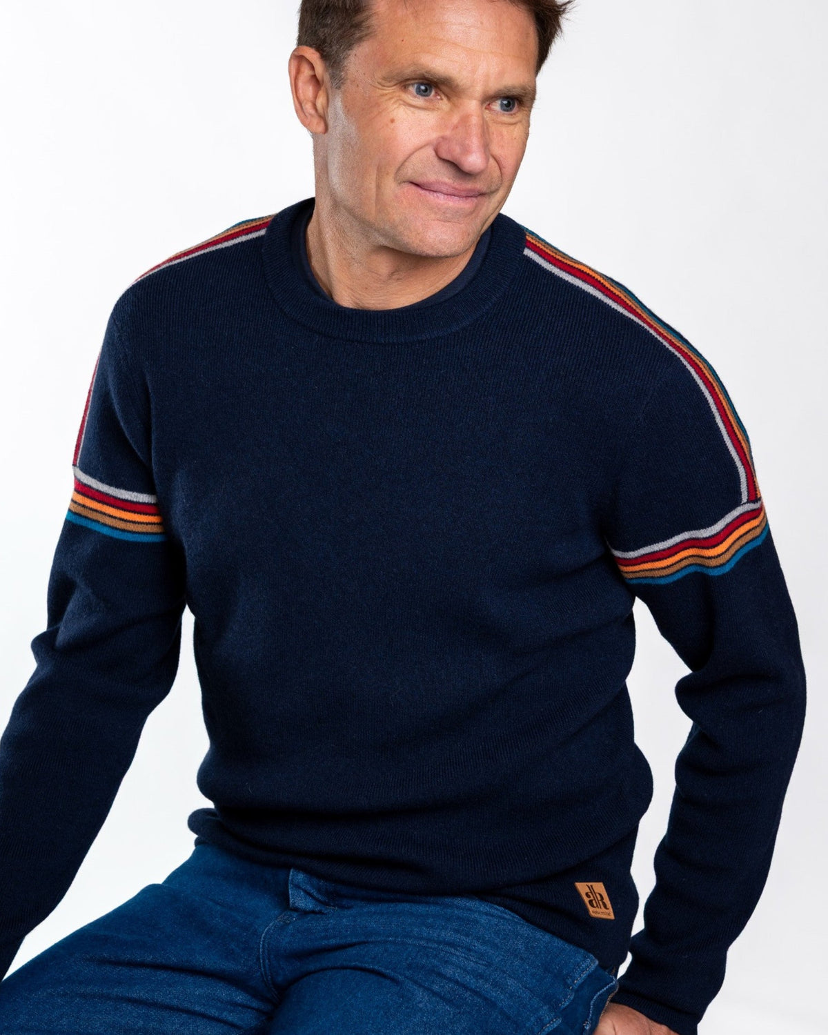 A man wearing a navy blue crewneck sweater with colorful striped details on the shoulders and sleeves, paired with blue jeans, seated against a plain white background. - Navy