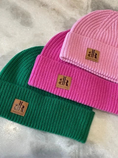 Three knit beanie hats in pink, green, and pink with a brand logo on a marble surface.