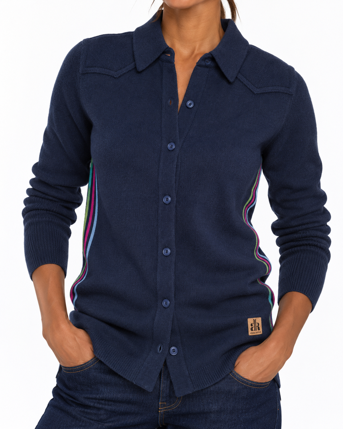 A woman wearing a navy blue knit button-up shirt with colorful striped accents along the sides and a small brown logo patch near the hem, paired with dark jeans against a plain white background. - Navy