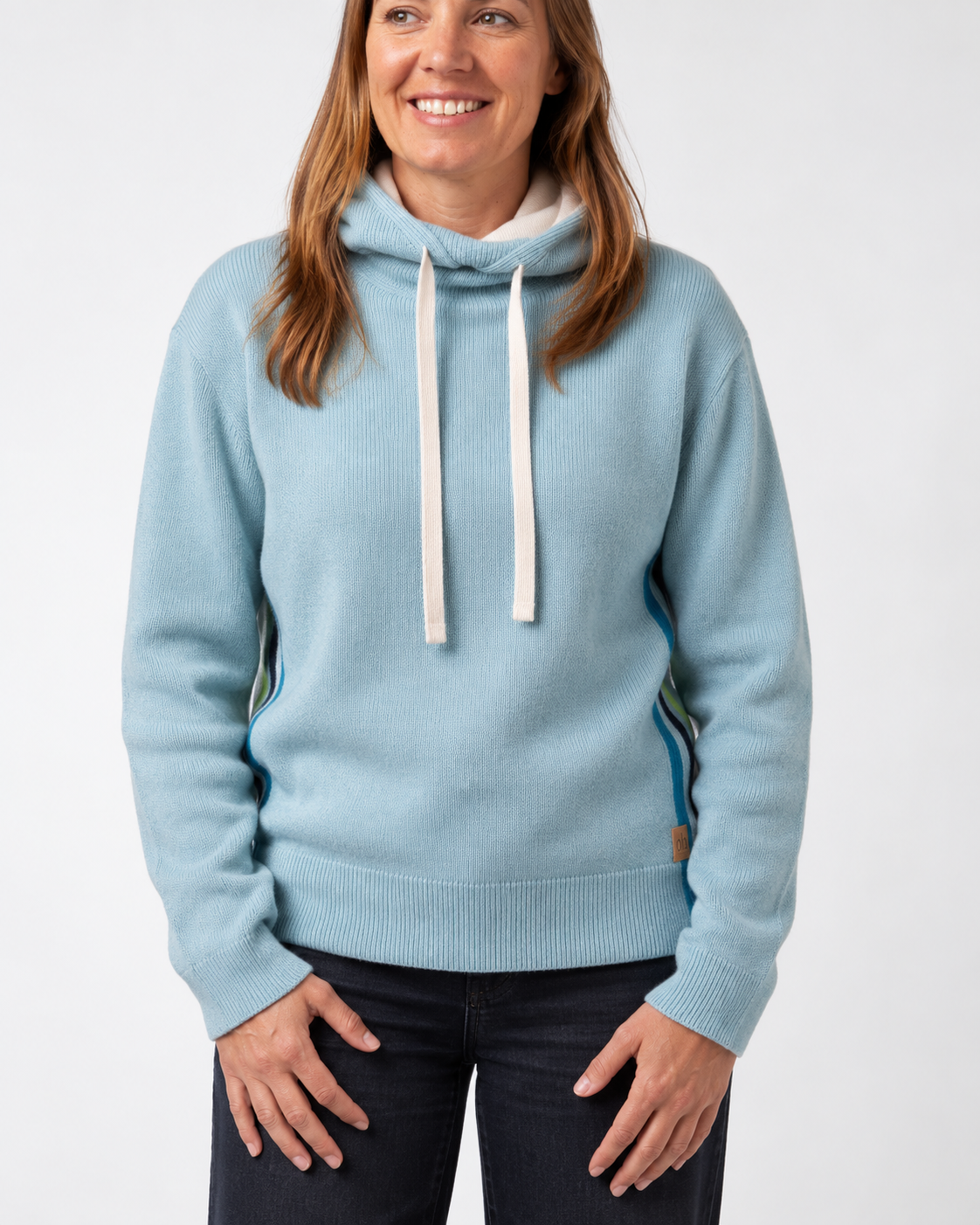 A woman wearing a light blue knit pullover hoodie with white drawstrings and subtle side stripe accents, paired with dark jeans, against a plain light gray studio background. - Sea