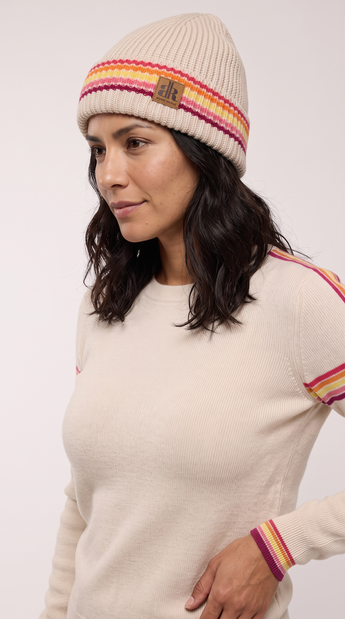 A woman wearing a cream ribbed knit beanie with pink, orange, and yellow stripes, paired with a matching cream sweater with striped details on the shoulders and cuff. - Ivory Stripes