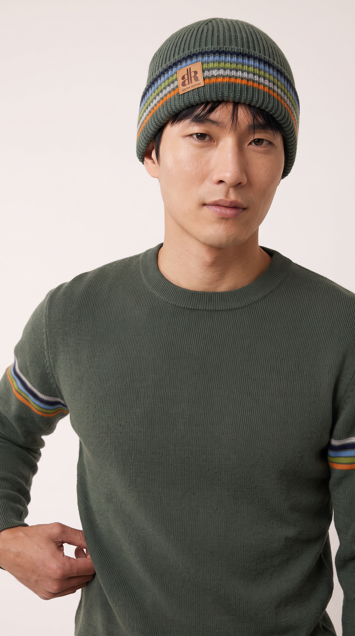 A man wearing a dark olive green knit crewneck sweater with striped accents on the sleeves and a matching striped knit beanie hat. The beanie features a small brown brand patch on the front. - Mountain Stripes
