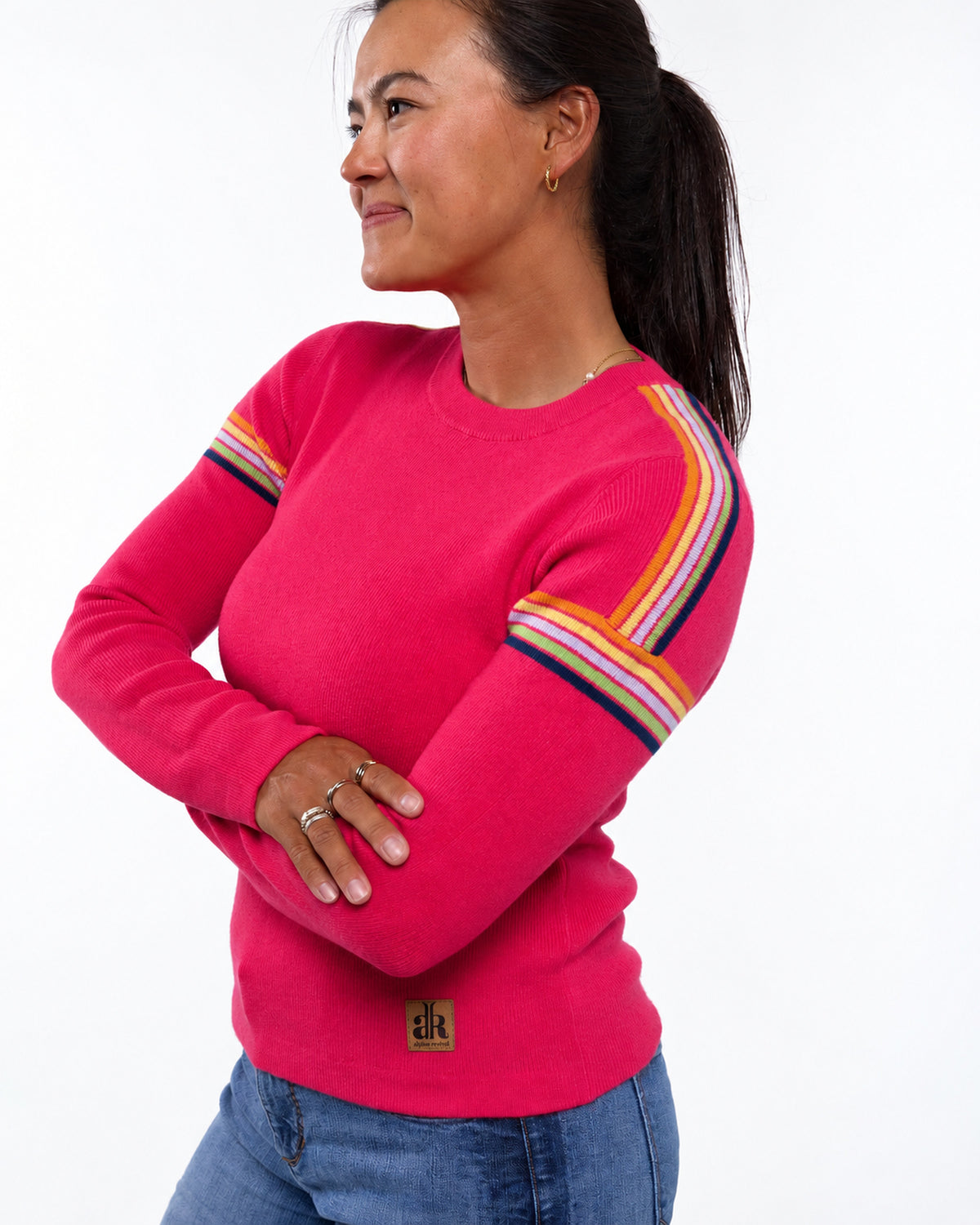 A woman wearing a bright pink crewneck sweater with colorful striped details on the sleeves, paired with blue jeans, photographed against a plain white background. - Watermelon